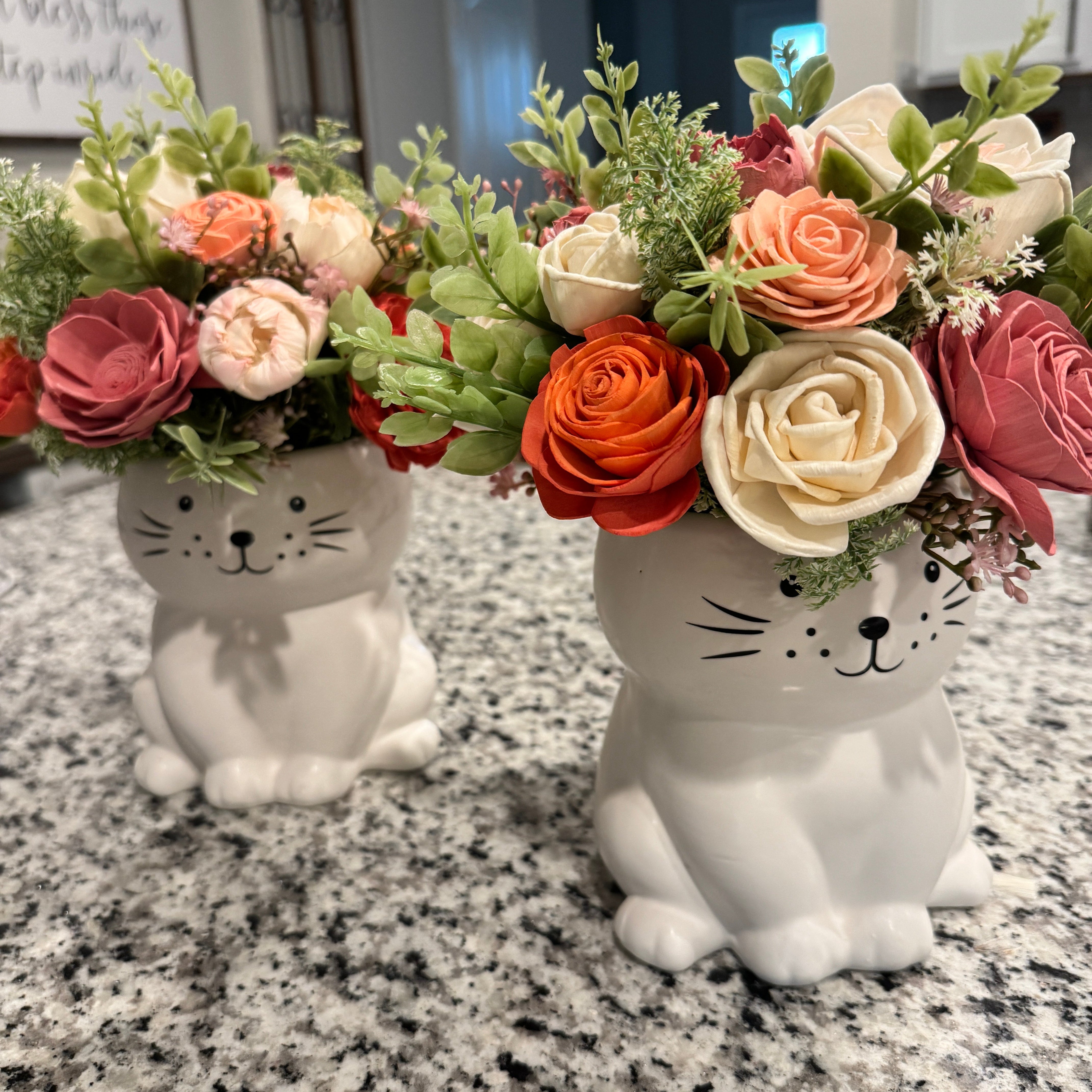 Two cat-shaped flower pots with floral arrangements on a granite countertop.