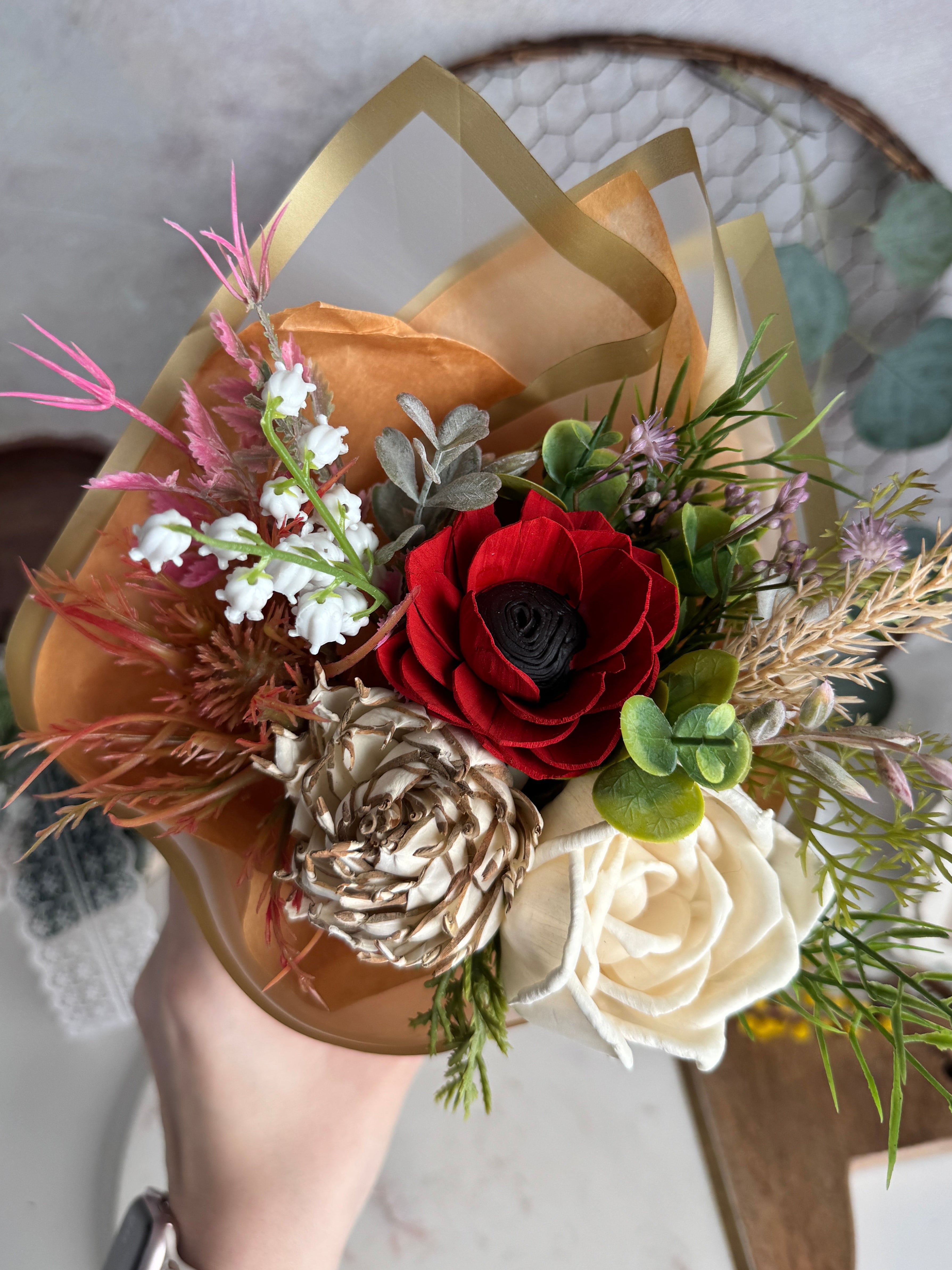 Hand holding a small bouquet with red flowers, white roses, and greenery against a neutral background.