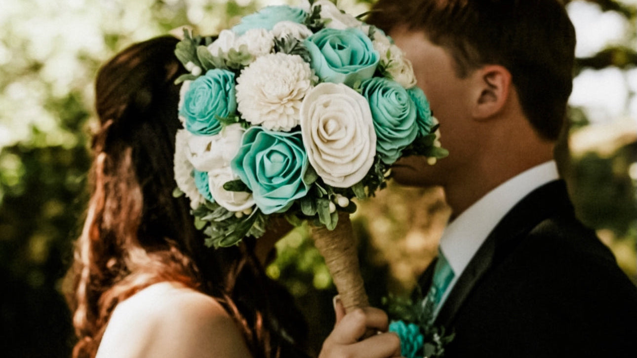 Bride holding a bouquet of turquoise and white flowers, standing next to a groom outdoors.