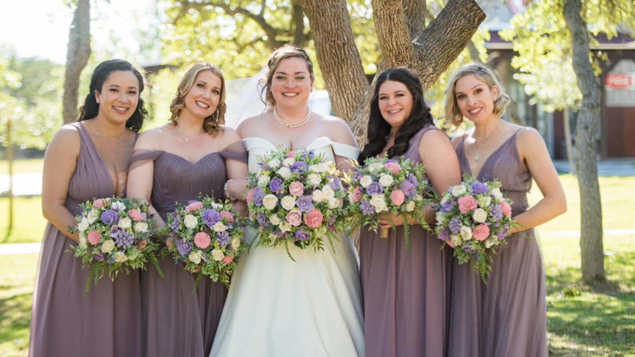 Bride with four bridesmaids in purple dresses holding floral bouquets outdoors.