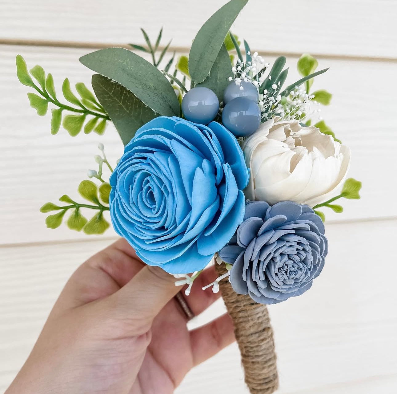 Hand holding a floral arrangement with blue, gray, and white flowers on a light wooden background