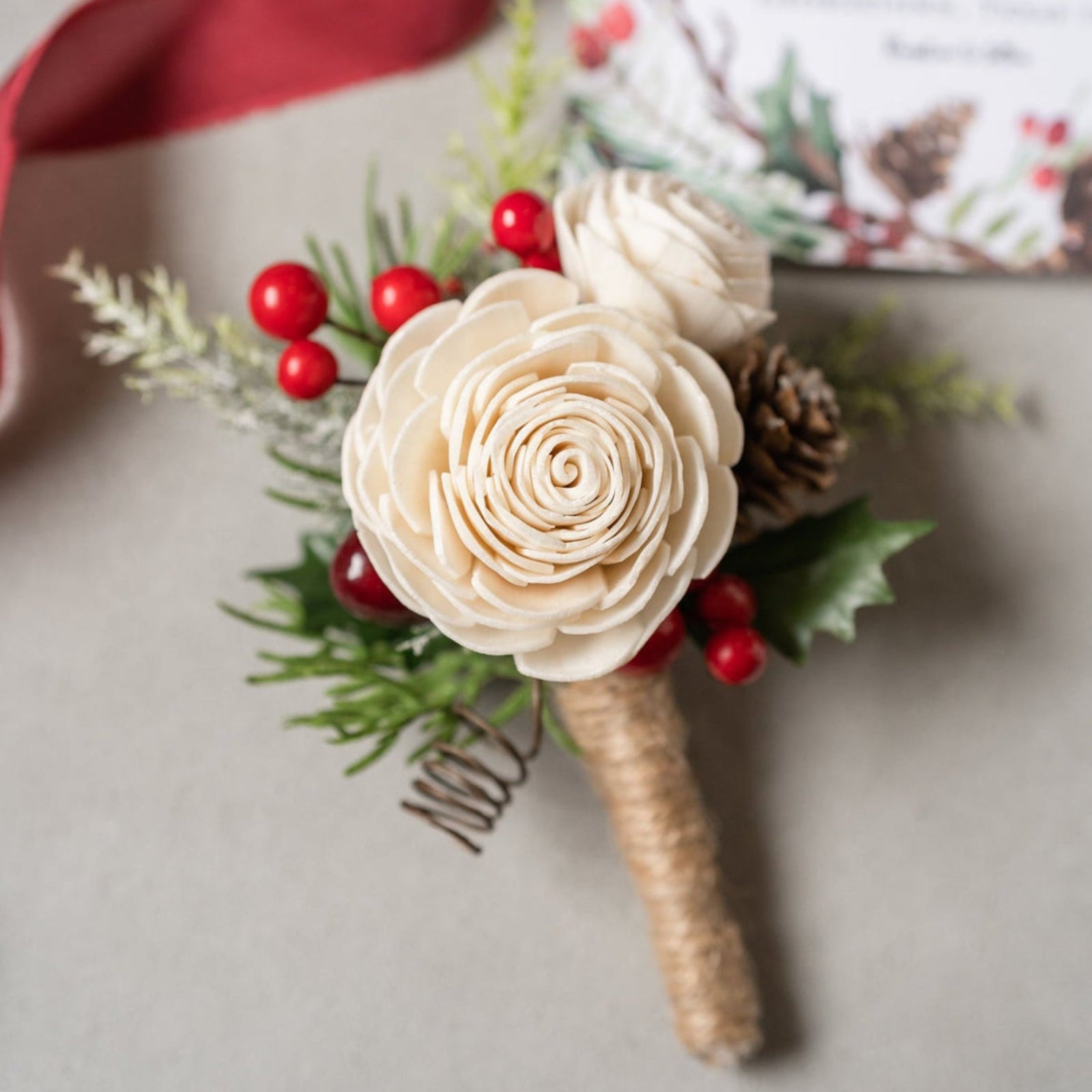 Christmas-themed boutonniere with beige flowers, red berries, and greenery on a light background.